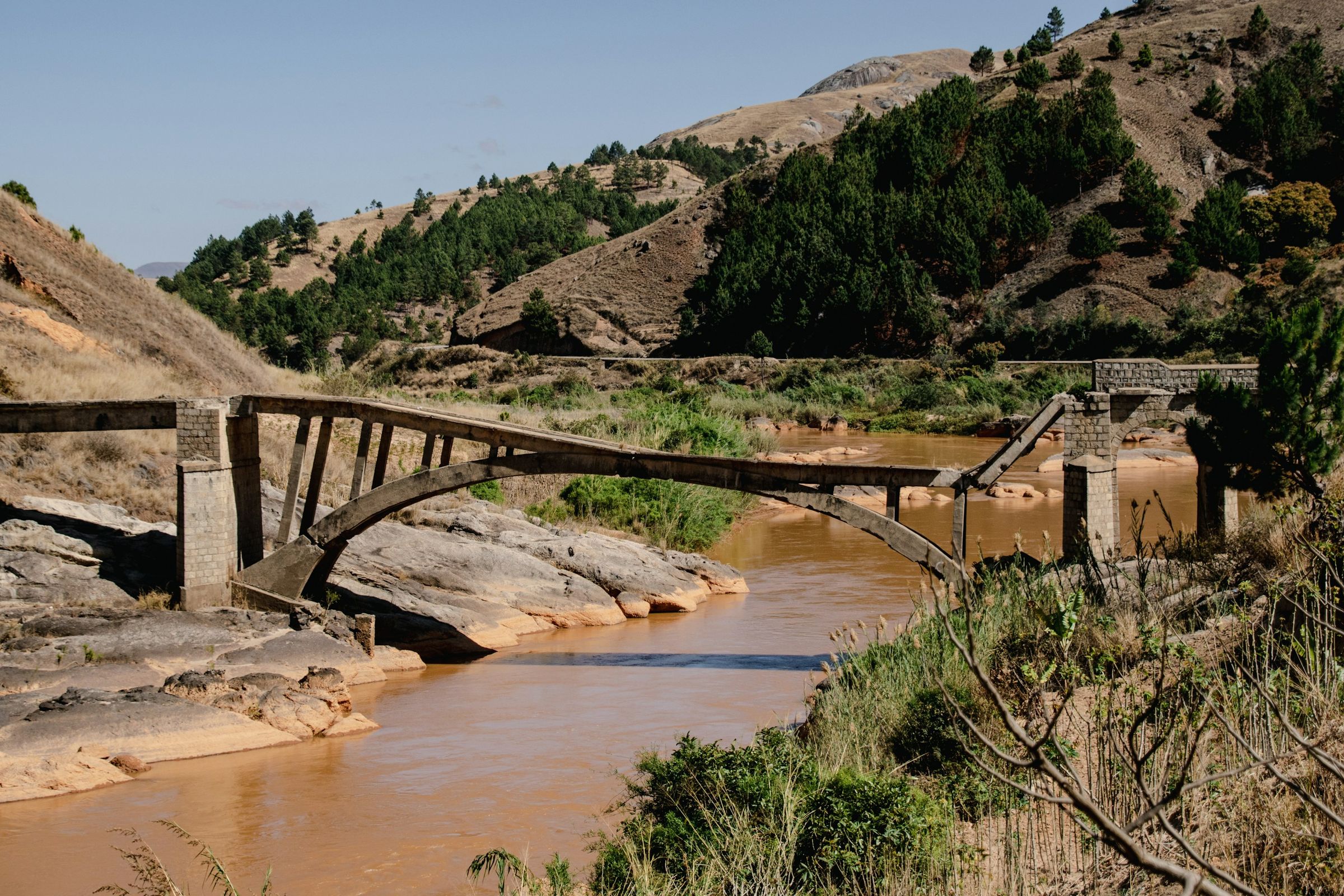 The broken arch of an old bridge over a muddy river beside the main road south